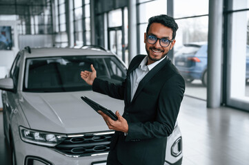Holding graphic tablet. Handsome indian man in suit is in the car showroom with the vehicle indoors