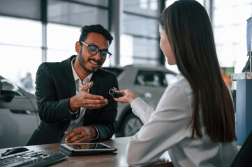 Woman is consulting man in the car showroom. Taking keys from the new automobile