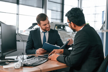 Sitting by the table. Two businessmen are working together in the car showroom