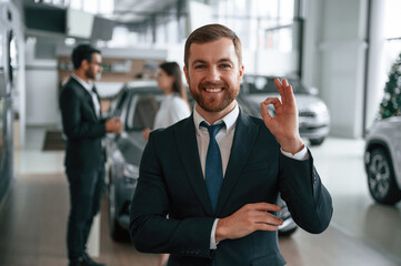 Handsome man is standing in front of his coworkers. Three people are together in the car showroom
