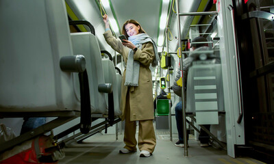 Long shot portrait of cute stylish woman standing and holding handrail in bus looking at smartphone smiling. Beautiful young caucasian female enjoying ride in public transport smiling.