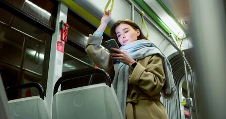 Low angle view of happy pretty young woman commuting by bus standing holding handrail. Attractive caucasian adult girl looking at smartphone checking social media smiling in empty trolleybus.