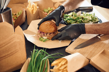 Close up view of burger, French fries and vegetables. Man is packing food into the paper eco boxes. Indoors, restaurant