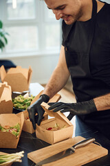 View from the side of meal, vegetables. Man is packing food into the paper eco boxes. Indoors, restaurant