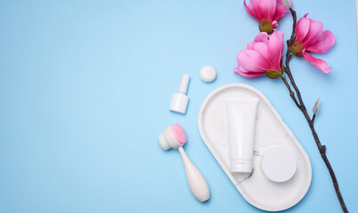 White plastic tubes and jars of cream, and a massage brush for facial cleansing on a blue background, items for cosmetic procedures