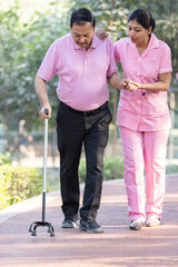 Female therapist supports senior man walking with cane at park.