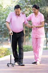 Female therapist supports senior man walking with cane at park.