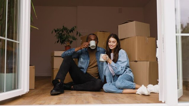 Happy african american man and caucasian woman moving to new home. Newly married couple sitting next to cardboard boxes and drinks coffee or tea
