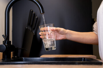 Close-up of a woman's hands pouring a glass of fresh water from the tap in the kitchen. Woman...