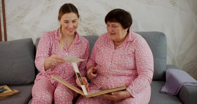 Senior granny and adult granddaughter looking at photos in family album together sitting on sofa at home wearing pink pijamas family look style. Memories, relationships, values, nostalgia concept.