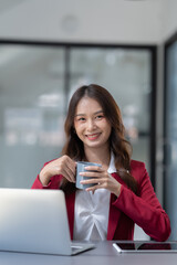 Beautiful Asian businesswoman holding a cup of coffee relax Take breaks and work hard on financial business matters at the office and have fun. Smiling happy working with the laptop.