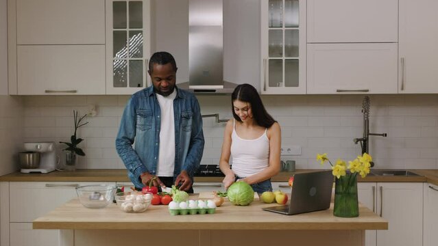 African american man and caucasian woman are cooking food together in the kitchen. The couple is talking and spending time together.