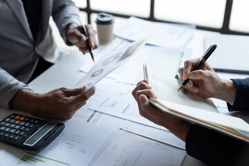 Two young Asian businessmen analyzing Calculate and discuss and brainstorm data, charts, graphs Taking notes on financial reports in notebooks at office financial advisors and accounting concepts.