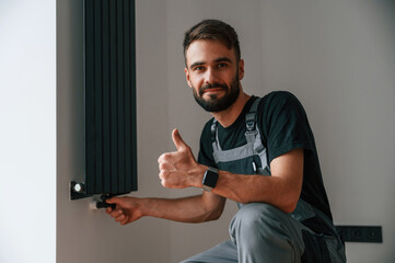 Sitting and showing thumb up. Plumber worker installing heating radiator in empty room of a newly...