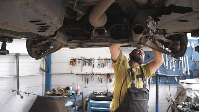 Professional bearded auto mechanic looks at camera and smile after finished work. Portrait of happy repairman repairing car at workshop. Positive man working underneath a lifting vehicle at garage