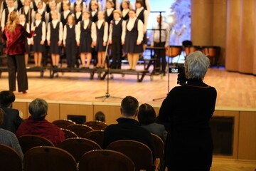 A person with a camera at a concert shoots a group of children with a conductor singing standing in...