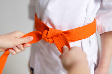 An adult's hands tie an orange karate belt on a child's kimono.