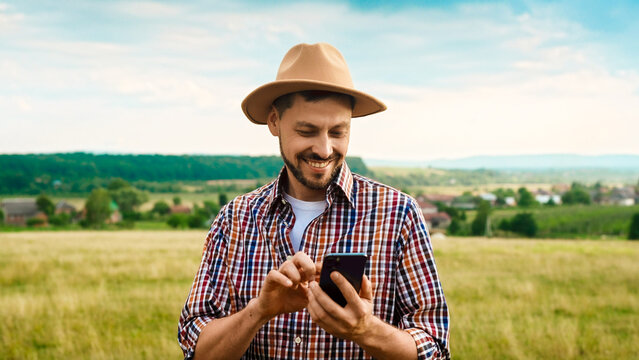 Caucasian handsome man in hat standing in grassland and using mobile phone. Outside. Good-looking young male farmer tapping and texting on smartphone. Gadget for farming. Chatting while working.