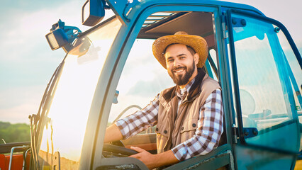 Portrait of Caucasian handsome happy young man farmer in hat sitting in cabin of tractor and smiling to camera. Countryside work concept. Good-looking male having rest from work at farm. Outdoors.