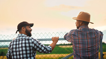 Rear on two young handsome Caucasian men farmers in hats standing at fence at farm on summer...