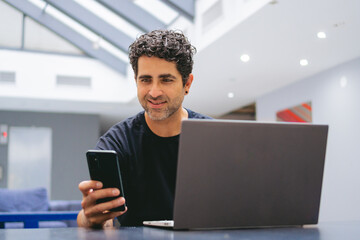 Middle-aged Latin man distracted by his cell phone instead of working on his laptop.