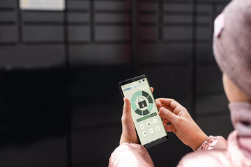 girl touch the button in the app at smartphone for opening box in modern postal automat. Self-service post terminal for storing parcels on the street.