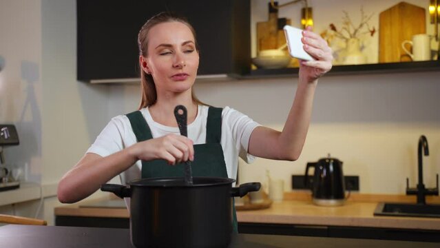 Joyful woman in an apron cooks at home and tastes a dish, shooting a video tutorial on the phone in the kitchen interior. Chef blogger cooks food according to a new delicious recipe. 