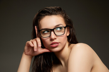 Portrait of beautiful brunette model girl with long straight hair and bare shoulders in eyeglasses over grey background. Concept of beauty, fashion, style, emotions