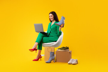 Happy woman with laptop and elegant shoes, consulting clients online, sitting on chair, yellow background, full length