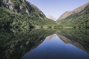 Naklejka na ściany i meble Morning landscape in the mountains with a lake in a valley between mountain slopes and rocks with a forest, a mirror surface of a mountain lake in the morning