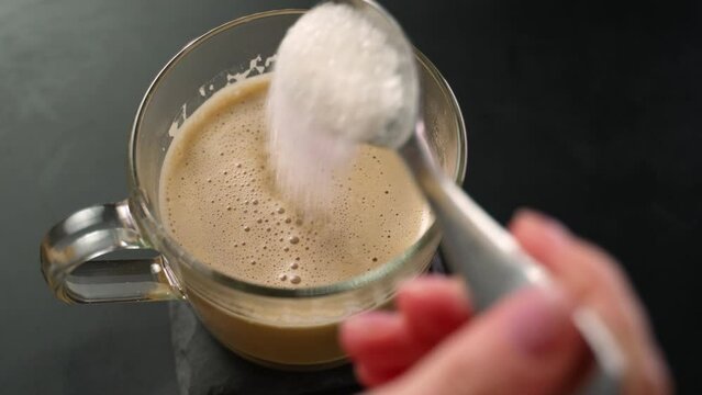 Woman's hand, with a coffee spoon, pours white sugar into a cup of coffee.
