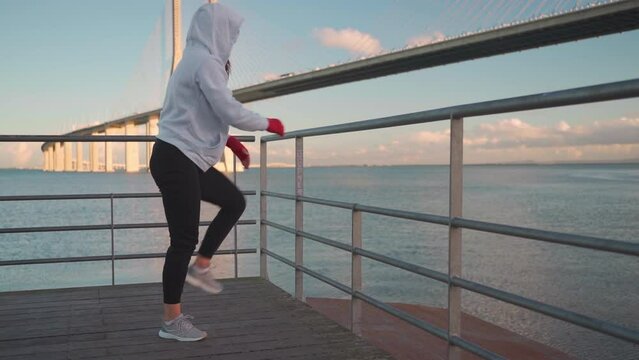 Athlete female doing push up and stratching exercises outdoor seascape background leaning on fence dressed hoodie sportswear. European athletic female practicing workout day. Slow motion full length