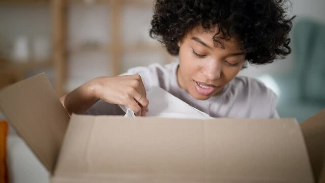 Happy amazed african american girl consumer holding opening cardboard box sit on sofa at home. Excited young woman receive unbox good parcel. Concept order postal shipping courier delivery. Unpacking.