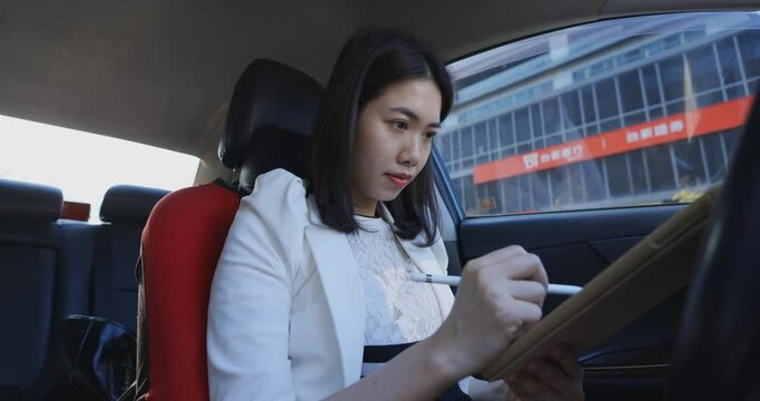 Asian businesswoman is using a tablet and a stylus inside the car to handle work tasks.	
