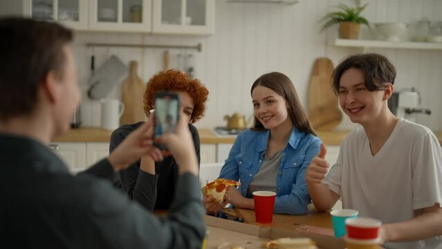 Friends gathered at home in kitchen, take joint photo. Guy takes pictures of beloved friends together. Men and girls sit at table, eat pizza, drink and enjoy talk. Happy leisure activity at weekend.