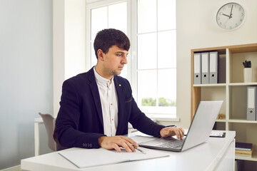 Fototapeta premium A young attractive business man working on a laptop at the office and looking on a computer screen and typing on the keyboard, considering new projects or startups, analyzing company.