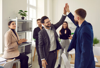 Two male colleagues doing high five at meeting in office. Office coworkers looking at them, smiling...