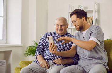 Young cheerful brunette nurse showing elderly man how to use smartphone. Nursing home service and patient support. Photo of caregiver teaching senior man to handle with mobile phone at home.