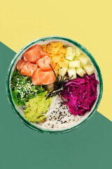 Bowl with salmon, avocado, red cabbage, nachos, cucumber, nori, white and black sesame, chuka. Round green bowl on a yellow and green background. Top view.