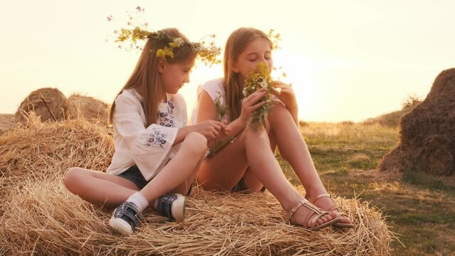 Two pretty sisters wearing wreath sitting on hay stack, sniffing flowers and weaving. Girls siblings spending time in the field in sunset time together. Beautiful female teens out of the city