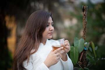 Young women holding a transparent tea cup with both hands with black tea in a garden wearing white woolen jacket