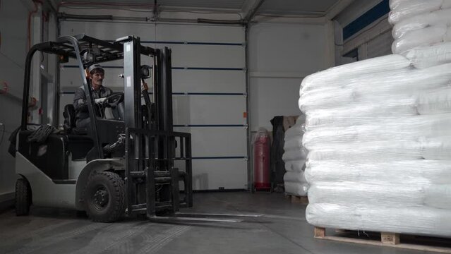 A worker in a warehouse unloads pallets with an electric forklift with sacks of flour. Adult Man works indoors on a forklift.