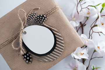 Empty egg-shaped gift tag on craft gift box on background with blooming apple tree branch. Flat lay, selective focus.