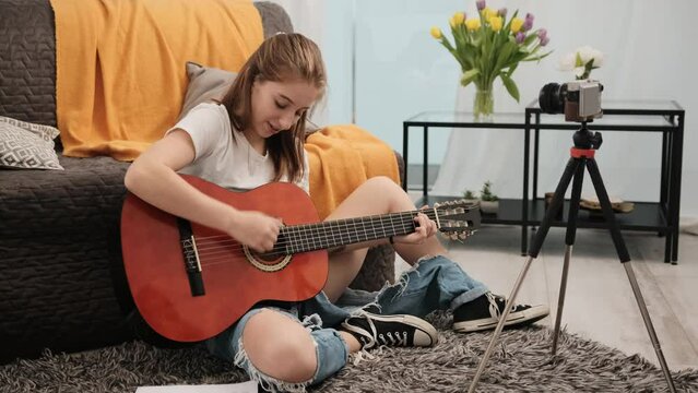 Young teenager girl with guitar making live stream to followers and explaining online how to play musical instrument. Pretty female guitaris making video on camera for social media channel