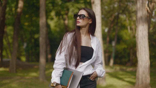 Portrait of beautiful young woman girl student in white shirt long-haired brunette in sunglasses holding books textbooks and standing in park near campus in sunlight