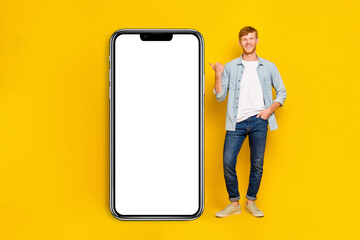 Photo of funny cool guy dressed denim shirt pointing thumb modern device empty space isolated yellow color background