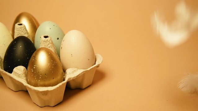 Easter is coming, a tray with painted eggs, on a nude background. Celebrations and traditions. Blue, gold and white with black egg, chicken feathers flying. Plastic eggs, place for text