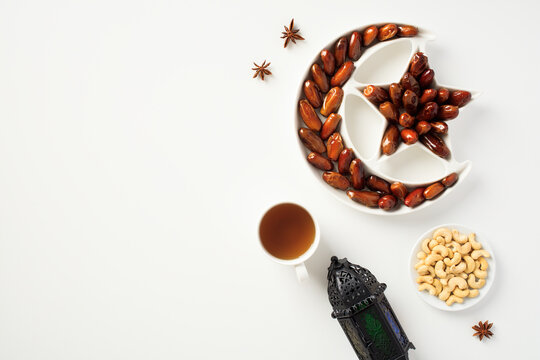Ramadan Kareem greeting card design. Dried dates in Islamic star and crescent moon plate, nuts, oriental lantern, cup of tea on white table. Flat lay, top view.