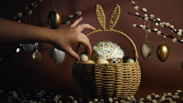 A woman's hand puts quail eggs in a basket in the shape of rabbit ears, with Easter cake and eggs. Happy easter and traditions, girl takes cute basket to church, place for text