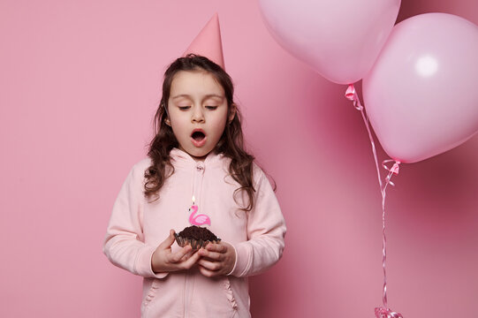 Lovely birthday girl in pink clothes and festive party hat, blowing out pinki flamingo candle on birthday cupcake, making cherished wish on isolated pink background with inflatable balloons